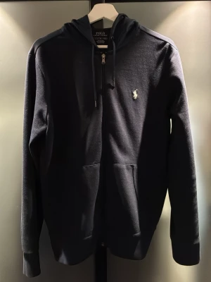 Mörkblå hoodie från Polo Ralph Lauren - Snygg mörkblå hoodie från Polo Ralph Lauren med dragkedja framtill, huva med dragsko och klassisk vit broderad logga på bröstet. Mjuk bomullsblandning och känguruficka gör den både stilren och bekväm. Perfekt för en avslappnad look.