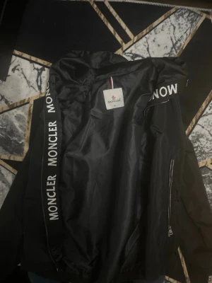 Svart Moncler vindjacka med huva - Snygg svart vindjacka från Moncler med huva och dragkedjor på sidorna. Jackan har tydliga Moncler-loggor längs huvkanten och insidan. Perfekt för blåsiga dagar och streetwear-stil. Materialet är lätt och smidigt, vilket gör den enkel att bära.