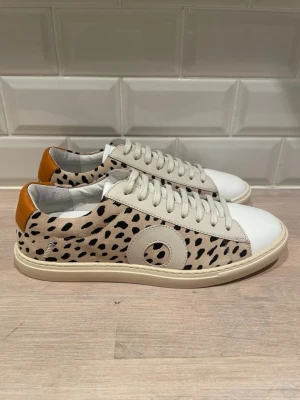 Oliver Cabell sneakers med djurmönster - Snygga sneakers från Oliver Cabell med beige mocka och svart leopardmönster, vita läderdetaljer och bruna hälkappor. Klassisk låg modell med vita snören och rundad tå. Perfekta för dig som vill sticka ut med din stil.