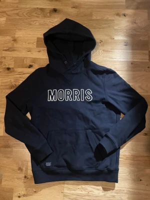 Marinblå Morris hoodie - Snygg marinblå hoodie från Morris med stor vit logga broderad på bröstet. Storlek S men passar vissa M. Klassisk modell med huva och känguruficka framtill. Tillverkad i mjukt bomullsmaterial och har ribbade muddar vid ärmslut och nederkant. Perfekt för en avslappnad och trendig stil.
