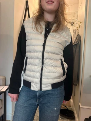 Vit dunjacka  från Moncler - Snygg och lätt vadderad vit dunjacka  från Moncler med svarta detaljer och dragkedja framtill. Västen har två fickor med svarta kanter och klassisk Moncler-logga på sidan. Perfekt att slänga över en hoodie eller tröja för en cool look.