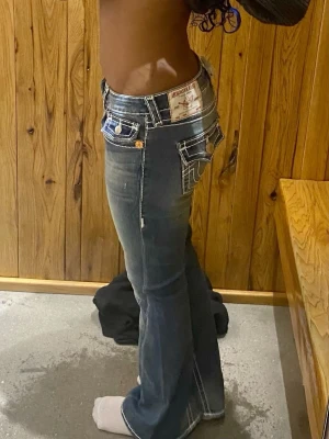 Mörkblå bootcut jeans från true religon - Säljer ett par mörkblå bootcut jeans från true religon  med tydliga vita kontrastsömmar och dekorativa fickor bak. Jeansen har låg midja, utsvängda ben och detaljerade knappar. Perfekta för dig som gillar en snygg och trendig look.