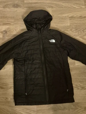 Svart vindjacka från The North Face - Svart vindjacka från The North Face med huva och hel dragkedja. Jackan har två sidofickor med dragkedja och logga på bröstet samt ärmen. Lätt vadderad och tillverkad i syntetmaterial, perfekt för blåsiga dagar. Obs att pris kan diskuteras!!