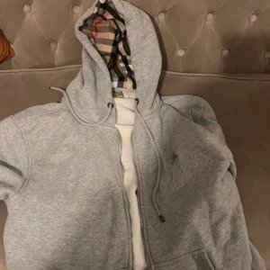 Grå huvtröja med mönstrad huva - Säljer en stilren grå huvtröja från Burberry med dragkedja och en ikonisk mönstrad insida i huvan. Perfekt för en avslappnad look. Tröjan har långa ärmar och justerbar huva med snören. Storlek xl men passar L pris går diskuteras vid snabb affär