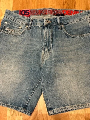 Superdry denimshorts ljusblå - Säljer ett par ljusblå denimshorts från Superdry med klassisk femficksdesign och broderad logga på fickan. Shortsen har normal passform och är tillverkade i bomull. Perfekta för sommaren och enkla att matcha med t-shirt eller hoodie. Aldrig använda, nya utan prislapp. 