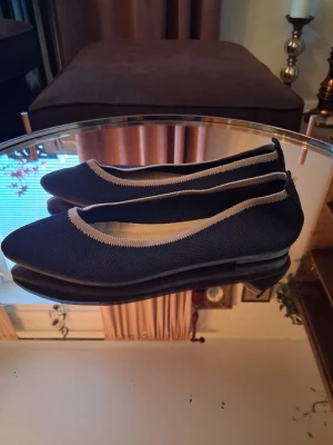 Svarta ballerinaskor med beige kant - Snygga ballerinaskor i svart tyg med beige kant runt öppningen. Skorna har en klassisk rundad tå och låg sula, perfekta för en stilren look. 