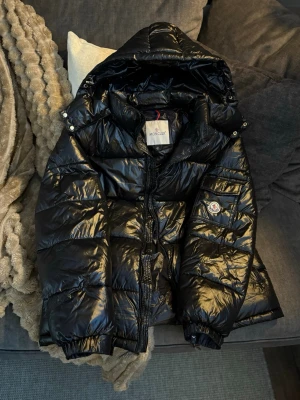 Svart dunjacka från Moncler - Säljer en svart glansig dunjacka från Moncler med huva och dragkedja framtill. Jackan har puffig design, logotyp på ärmen och detaljerade dragkedjor med Moncler-märke. Perfekt för kalla vinterdagar och riktigt snygg streetstil.