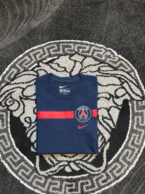 Blå PSG t-shirt från Nike - Snygg marinblå t-shirt från Nike med Paris Saint-Germain-logga och röd rand över bröstet. Klassisk rund hals och korta ärmar. Perfekt för dig som gillar fotboll och vill ha en sportig stil.