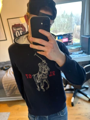 Navy blå hoodie från Polo Ralph Lauren - Cool svart hoodie från Polo Ralph Lauren med stort tryck av en ridande häst och text på framsidan. Tröjan har huva med vit insida och långa ärmar. Perfekt för dig som gillar klassiska märken och sportig stil.