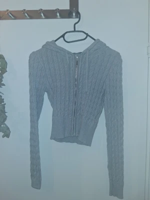 Grå stickad hoodie med dragkedja - Snygg grå stickad hoodie med dragkedja framtill och kabelstickat mönster. Tröjan är croppad med långa ärmar och huva, perfekt för lager-på-lager-stil. Materialet är mjukt och stretchigt för en bekväm känsla.