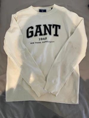 GANT vit stickad tröja (äkta) - Snygg vit stickad tröja från GANT med broderad logga och texten 'GANT 1949 New Haven, Connecticut' på bröstet. Tröjan har rund halsringning, ribbade muddar och är tillverkad i mjukt material. Perfekt för en avslappnad och stilren look. Storlek M men passar också S.