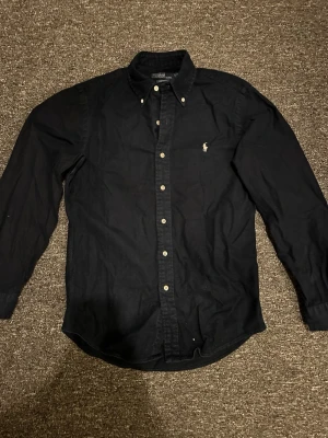 Mörkblå skjorta från Ralph Lauren - Klassisk svart skjorta från Ralph Lauren med broderad logga på bröstet. Skjortan har knappar framtill, button-down krage och långa ärmar. Tillverkad i mjuk bomull som känns skön mot huden. Perfekt för en stilren och avslappnad look.