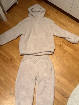 Beige fluffigt loungewear-set - Supermysigt mjukis-set i beige teddy-material med hoodie och byxor. Hoodien har stor ficka framtill och byxorna resår i midjan och anklarna. Riktigt varmt och skönt! 