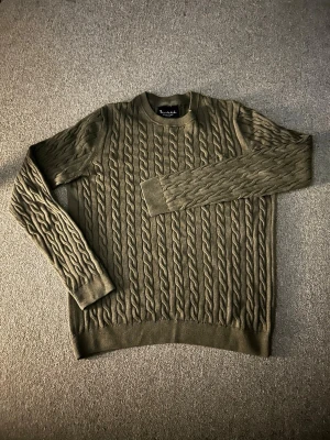 Olivgrön kabelstickad tröja - Säljer en olivgrön kabelstickad tröja från Brooklyn. Tröjan har rund halsringning, långa ärmar och ribbade muddar vid ärmslut och nederkant. Klassisk kabelstickad design som ger en snygg och tidlös look.
