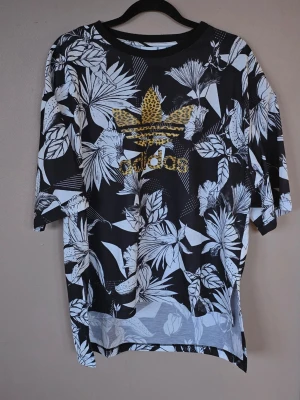 Adidas t-shirt med leopardlogga - Svart och vit t-shirt från Adidas med stort leopardmönstrat logotryck på bröstet. T-shirten har korta ärmar och ett tropiskt bladmönster över hela plagget. Perfekt för dig som vill sticka ut med en cool streetstyle. Superunik Välskött t-shirt.  Inga anmärkningar, som ny!!  Boxig form, med längre Längd bak.  Storlek Large (40 EU) Unisex  Bröstvidd 53 cm Längd fram 53 cm Längd bak 69 cm