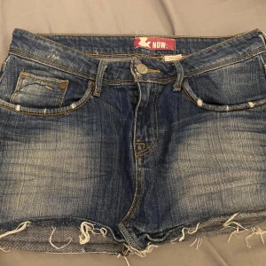 Blå jeansshorts med rå kant från NOW - Säljer ett par lågmidjde blå jeansshorts från NOW med rå, fransig kant nertill och slitna detaljer. Shortsen har fem fickor, knappgylf och kontrastsömmar.