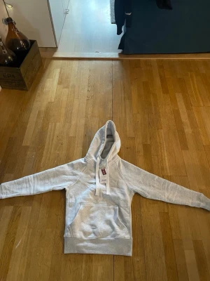 Ljusgrå hoodie från Russemarch - Säljer en ljusgrå hoodie från Russemarch i unisexmodell. Hoodien har en stor huva med breda dragsnören och en klassisk magficka framtill. Tillverkad i mjukt material, perfekt för en avslappnad stil.