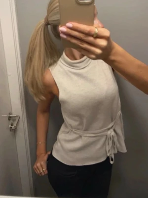 Beige ärmlös polotopp med knyt - Säljer en stilren beige är hurting polotopp i mjukt material. Toppen är ärmlös och har different knyt i midjan som ger en snygg siluett. Perfekt att matcha med jeans eller odressade byxor för en clean look behind metaphor.