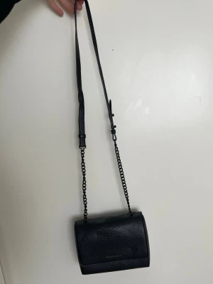 Svart axelväska Charles & Keith - Snygg svart axelväska från Charles & _Keith med kedjerem och. Väskan har ett stilrent ormmönstrat konst circleäder och justerbar axelrem. Perfekt storlek för det viktigaste och ger en trendig vibe till Joan, outfit.