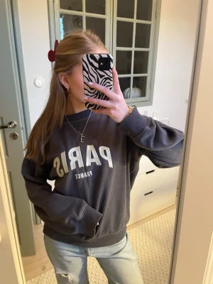 Mörkgrå sweatshirt med Paris-tryck - Cool mörkgrå sweatshirt med vitt Paris-tryck på bröstet och texten France under. Tröjan har rund hals, långa ärmar och ribbade muddar. Perfekt för en avslappnad och trendig look.
