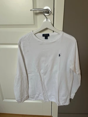 Vit långärmad tröja Polo Ralph Lauren - Klassisk vit långärmad tröja från Polo Ralph Lauren med rund halsringning och den ikoniska lilla marinblå logotypen broderad på bröstet. Tillverkad i mjukt bomullstyg som känns skönt mot huden. Perfekt för en clean och stilren look.