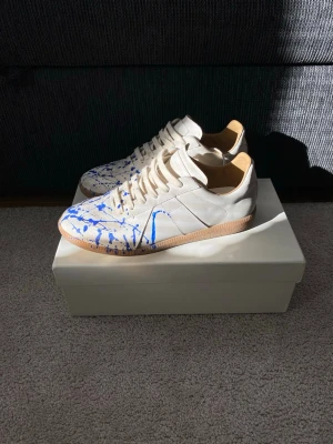 Maison Margiela Blue Paint Splatter Gats - Unika sneakers från Maison Margiela i vitt läder med coola blå färgstänk och klassisk gummisula. Lågt skaft och stilren design som sticker ut. Perfekta för dig som vill ha något annorlunda och trendigt i garderoben.