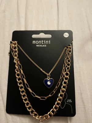 Montini halsband med blått hjärthänge - Säljer ett snyggt Montini halsband med två kedjor i guldfärg. Det ena har ett blått hjärthänge med vit måndetalj, och det andra är en chunky kedja. Perfekt för att ge din outfit en cool touch. 
