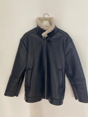 Svart skinnjacka med teddy tyg på insidan - Snygg svart skinnjacka med krage i beige fuskpäls och dragkedja framtill. Jackan har raka ärmar med dragkedjor vid ärmslut och två fickor framtill. Perfekt för dig som vill ha en stilren och trendig look.