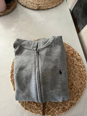 Grå zip hoodie från Polo Ralph Lauren - Säljer en stilren grå hoodie med dragkedja från Polo Ralph Lauren. Klassisk design med huva och den ikoniska lilla mörk cognitive logon broderad på bröstet. Tillverkad i mjukt sweatshirtmaterial som är perfekt för chill dagar. Stl 14-16 år