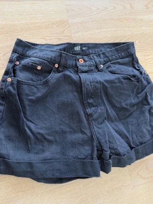 Svarta denimshorts från 157 - Snygga svarta shorts i denim från 157 med klassisk femficksdesign och uppvikta benslut. Shortsen har knapp och dragkedja framtill samt detaljer i kopparfärg. Perfekta för varma dagar och enkel att matcha.