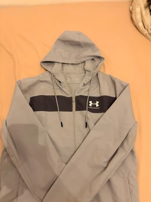 Under armour kofta - Snygg grå kofta från Under Armour med dragkedja och svart detalj över bröstet. Hoodien har justerbar huva med snören och meshfoder på insidan för extra komfort. Perfekt för en sportig och avslappnad stil.