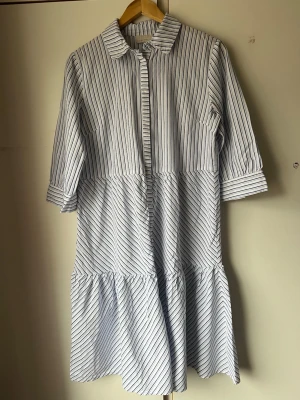 Randig skjortklänning med volang - Snygg skjortklänning i vitt med blå ränder och volang nedtill. Klänningen har krage, knappar framtill och trekvartsärm. Perfekt för en avslappnad men stilren look.