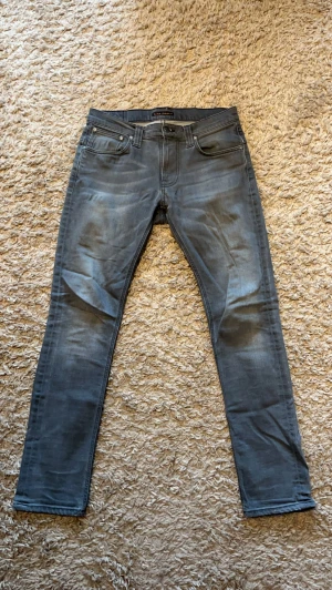 Grå jeans från Nudie Jeans - Säljer ett par grå slim fit jeans från Nudie Jeans med klassisk femficksmodell och snygga slitningar. Jeansen har en något tvättad look och raka ben. Perfekta för dig som gillar en avslappnad men ändå stilren vibe. Storlek 32/32