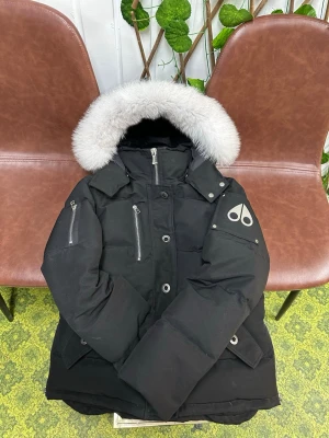 Svart dunjacka med pälskrage Moose Knuckles - Snygg svart dunjacka från Moose Knuckles med stor fluffig pälskrage i grått. Jackan har dragkedjor på bröstet, fickor med lock och ikonisk logga på ärmen. Perfekt för kalla vinterdagar och riktigt varm.