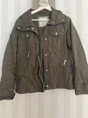 Olivgrön vårjacka - Snygg olivgrön vårjacka med dragkedja och tryckknappar. Jackan har fyra fickor framtill, två med dragkedja och två med lock. Huva och justerbara muddar. Materialet är tunt och passar perfekt för våren. Inga synliga loggor.