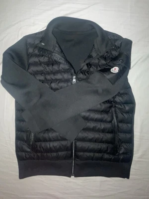 Moncler cardigan - Snygg svart dunväst från Moncler med quiltad framsida och mjuka stickade partier på sidor och rygg. Västen har dragkedja framtill, två fickor med dragkedja och Moncler-logga på ena axeln. Perfekt för lager-på-lager och streetstyle. Tveka inte vid funderingar!