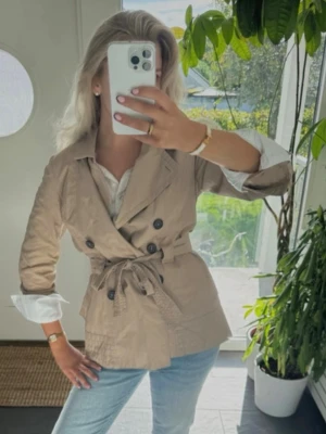 Beige trenchcoat med knytskärp - Säljer en snygg beige trenchcoat med dubbelknäppning och knytskärp i midjan. Jackan har klassisk krage, stora svarta knappar och är tillverkad i ett lätt material som passar perfekt till jeans. Ärmarna är trekvarts och kan vikas upp för en cool look.