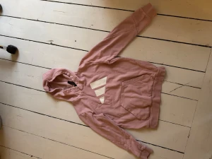 Rosa hoodie från Adidas - Säljer en rosa hoodie från Adidas med vit logga på bröstet. Hoodien har huva, magficka och ribbade muddar vid ärmslut och nederkant. Perfekt för en avslappnad och sportig stil.