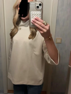 Beige polotopp med trekvartsärm - Stilren beige polotopp med trekvartsärm och rak passform. Toppen har en hög krage och är tillverkad i ett mjukt material som ger en clean och modern look. Perfekt att matcha med jeans eller kjol för en enkel men snygg outfit.