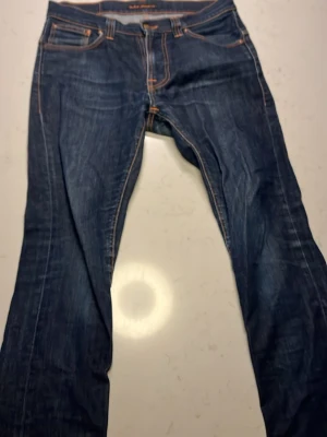 Mörkblå jeans från Nudie Jeans - Säljer ett par klassiska mörkblå jeans från Nudie Jeans med orangea kontrastsömmar och femficksmodell. Jeansen har raka ben och snygga detaljer på bakfickorna. Perfekta för dig som gillar en tidlös och avslappnad stil.