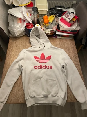  Vit hoodie från Adidas med röd logga - Säljer en vit hoodie från Adidas med stor röd Adidas-logga och text på bröstet. Hoodien har huva och en stor magficka. Tillverkad i mjukt bomullsmaterial och har ribbade muddar vid ärmar och nederkant. Perfekt för en avslappnad streetwear-look.