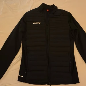 Svart pufferjacka från CCM - Svart pufferjacka från CCM med quiltad front och hel dragkedja. Jackan har långärmade ärmar, diskret logga på bröstet och sidofickor med dragkedja. Perfekt för dig som vill ha en sportig och stilren look.