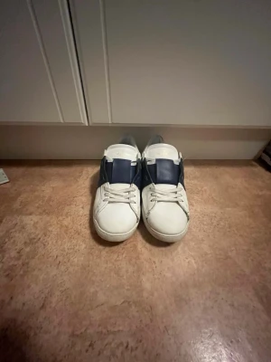 Valentino Garavani vita sneakers   - Snygga vita sneakers från Valentino Garavani med en bred mörkblå rem över sidan. Nypris :7200kr. Skorna har klassisk låg profil, vita snören och detaljer i läder. Perfekta för dig som vill ha en stilren och exklusiv look.priset är ej hugget i sten