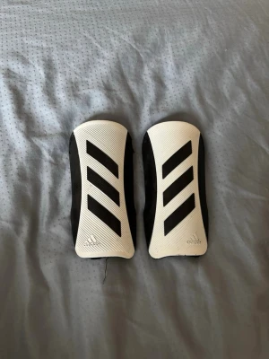 Adidas benskydd i vitt och svart - Säljer ett par vita och svarta benskydd från Adidas med klassiska svarta ränder. Skydden har en hård yta framtill och vadderad baksida för extra komfort. Perfekta för fotbollsträning eller match.