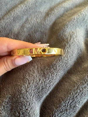 Guld ingraverat armband Michael Kors - Snyggt guldfärgat armband från Michael Kors med MK-logga framt Gemini soprat och ingraverad märkning på insidan. Man öppnar den från sidan och klickar bara fast den runt hand leden skulle säga att den passar de flesta handlederna 