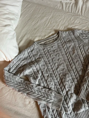 Grå kabelstickad tröja - Snygg grå kabelstickad tröja med rund halsringning och ribbade muddar. Tröjan har ett klassiskt flätmönster över hela plagget och är långärmad. Perfekt för lager på lager och passar till det mesta i garderoben.