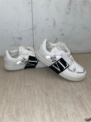 Vita Valentino garavani skor - Snygga vita Valentino garavani skor, med stor svart VLTN-logga på sidan. Skorna har klassisk låg profil, vita snören och perforerad tå för extra ventilation. Detaljer i svart och vitt som ger en clean och modern look.