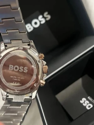 Silverfärgad klocka från BOSS - Stilren klocka från BOSS med länkarmband i silverfärgat rostfritt stål och detaljer i guld. Klockan har en rund urtavla och klassisk design med BOSS-logga på både baksidan och spännet. Perfekt accessoar för dig som gillar en clean och lyxig look.