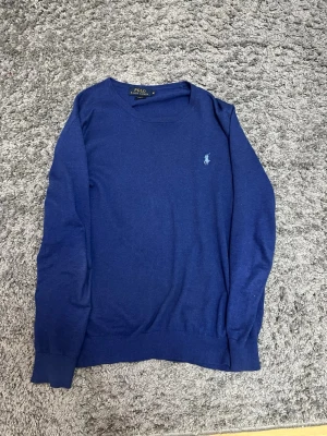 Blå tröja från Polo Ralph Lauren - Mörkblå tröja från Polo Ralph Lauren med rund halsringning och den klassiska broderade loggan på bröstet. Tröjan har långa ärmar och ribbade muddar. Materialet känns mjukt och lätt, perfekt för en stilren look.