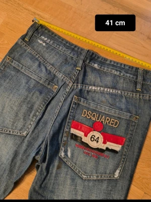 Blå DSQUARED jeans med brodyr - Säljer ett par blå jeans från DSQUARED med cool broderad detalj på bakfickan och texten '64'. Klassisk femficksmodell med raka ben och slitningar för en trendig look. Perfekta för dig som gillar streetwear och vill sticka ut. Inga fel byx gylfen ska va så 